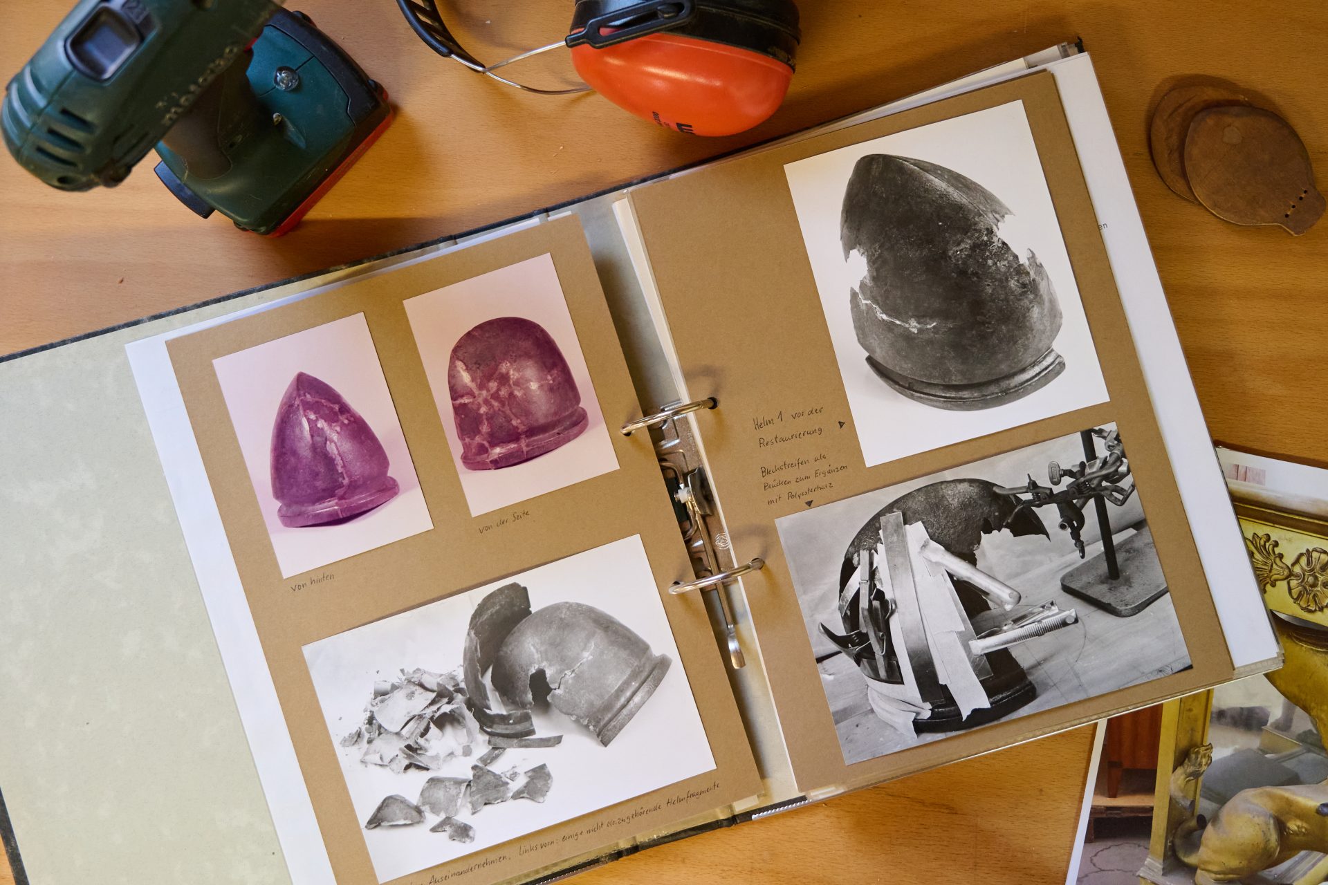 Ein geöffnetes Album mit Fotos von Hüten und Ausstellungsstücken, umgeben von Werkzeugen und Gehörschutz.