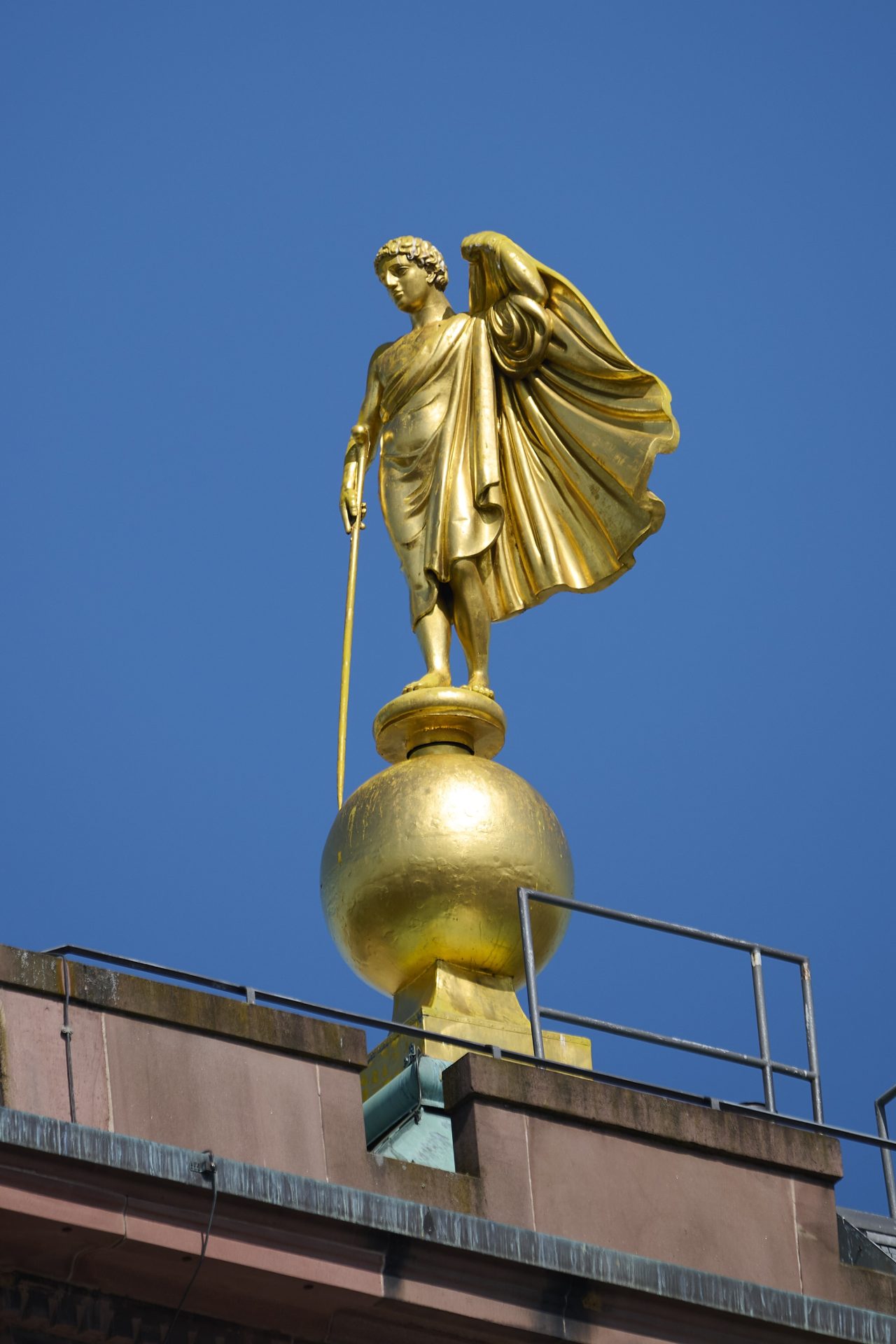 Goldene Statue mit Umhang auf einer Kugel vor klarem blauen Himmel.