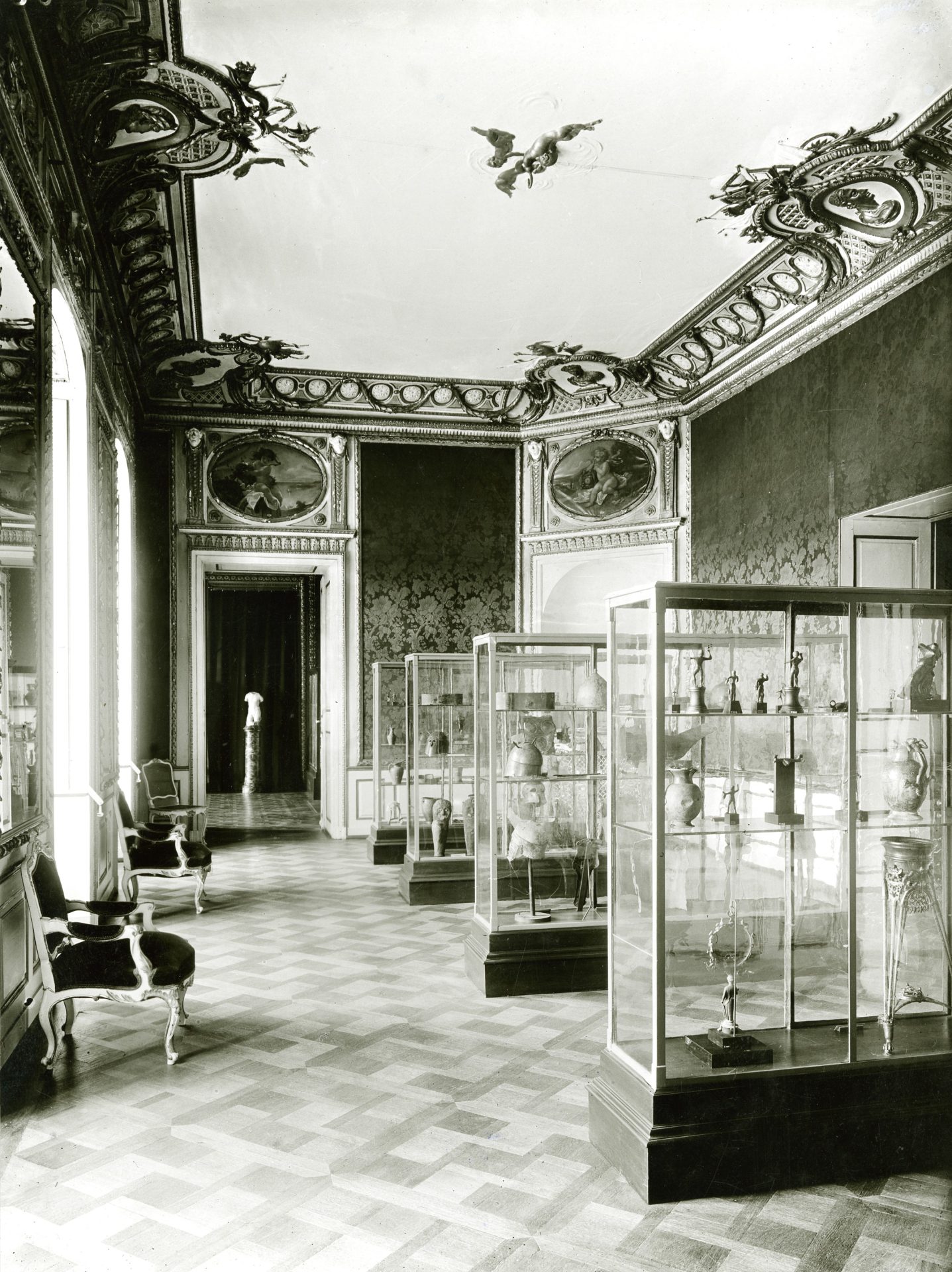 Historischer Raum mit kunstvoller Decke, Glasvitrinen und antiken Exponaten. Elegante Möbel im Vordergrund.