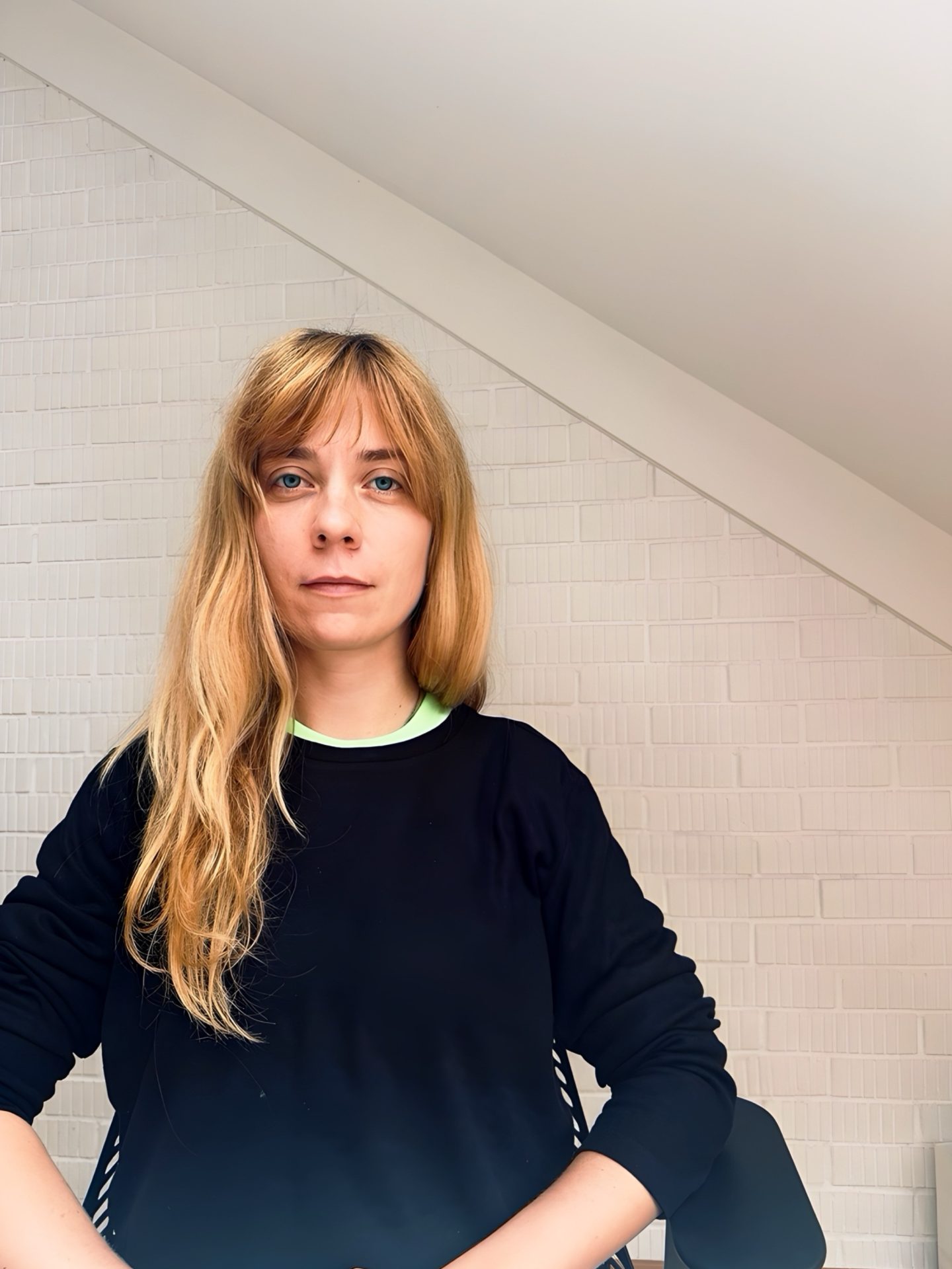Frau mit langen blonden Haaren sitzt vor einer hellen Wand und blickt direkt in die Kamera. Sie trägt einen schwarzen Pullover.