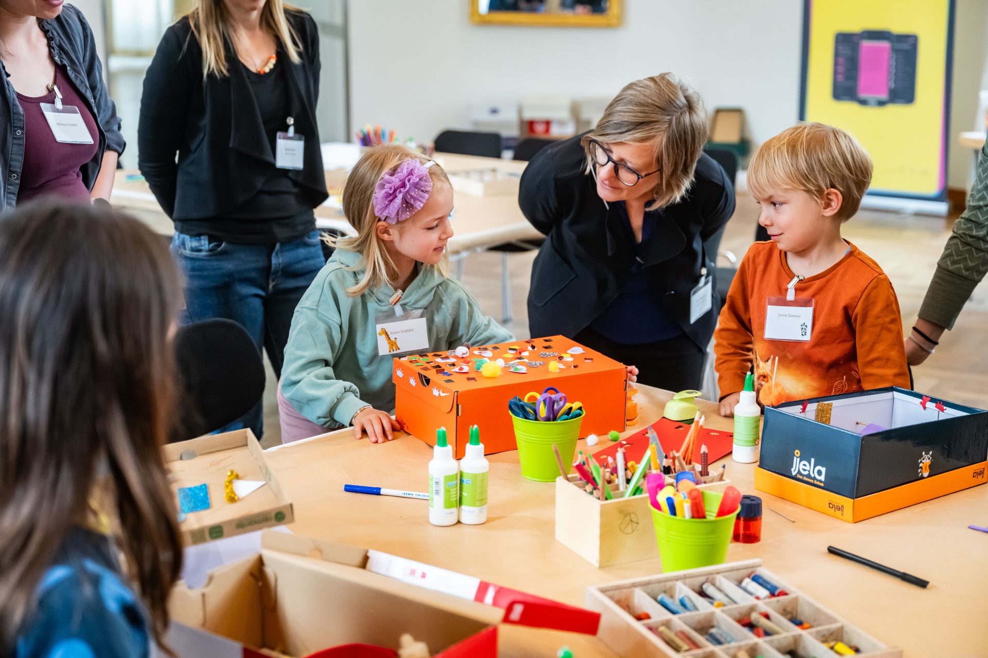 Kinder und Erwachsene gestalten gemeinsam farbenfrohe Projekte in einem kreativen Raum mit sichtbaren Stiften und Materialien.
