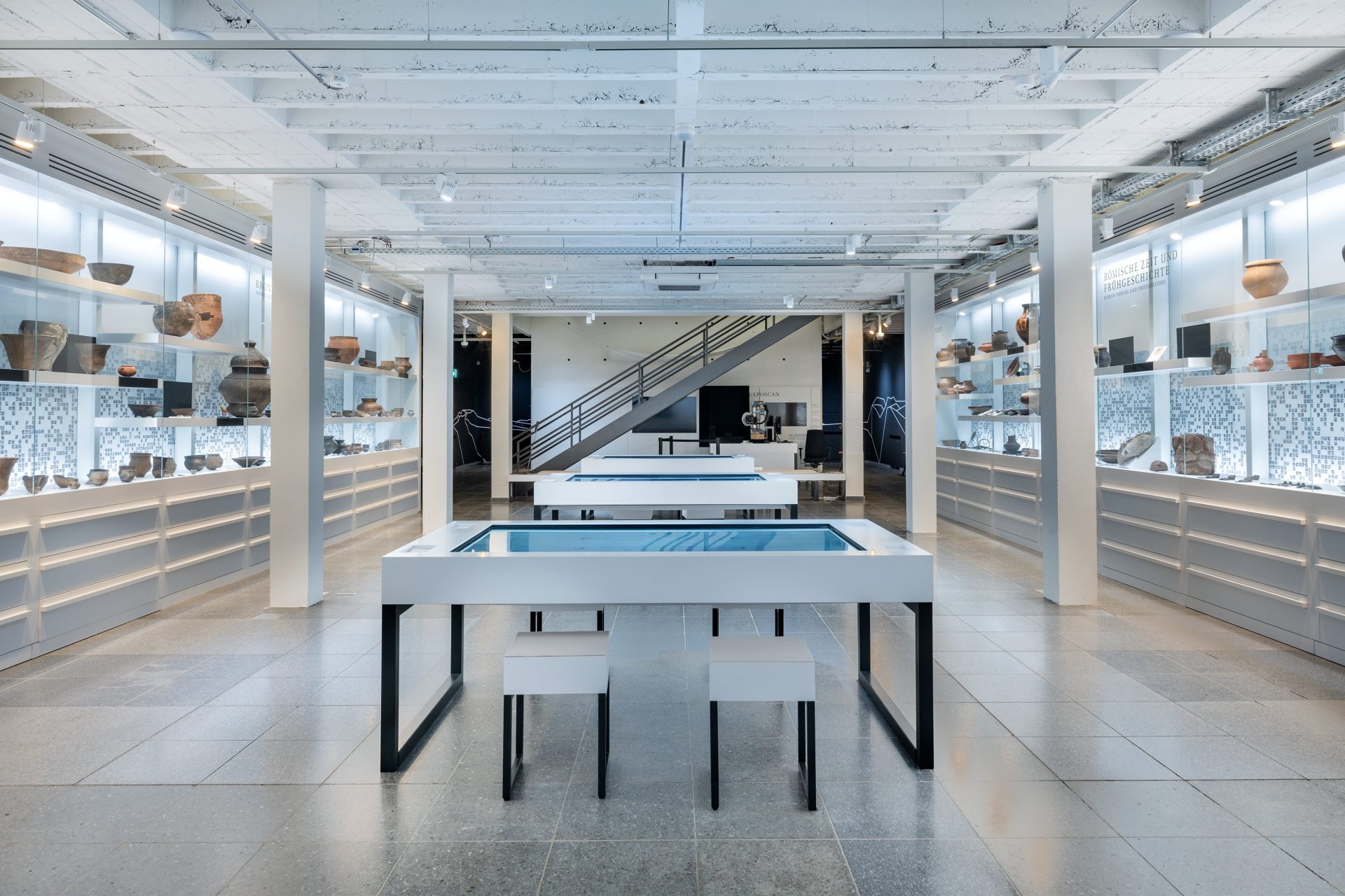 Moderner Ausstellungsraum mit Tischen, Vitrinen und historischen Artefakten im Hintergrund. Helle, offene Atmosphäre.