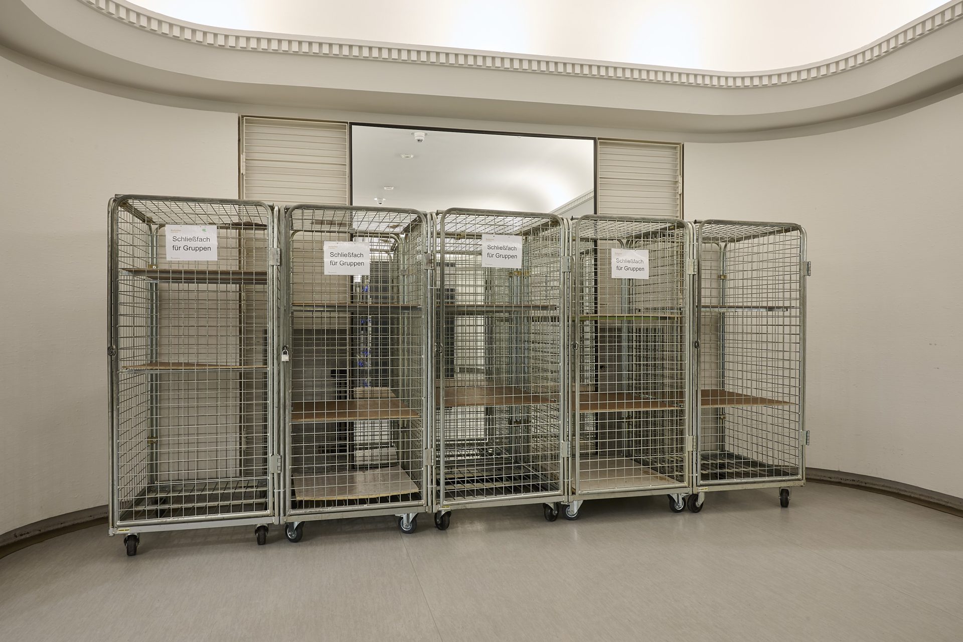 Vier Rollenkäfige in einem hellen Raum, beschriftet: "Gruppenschließfach".