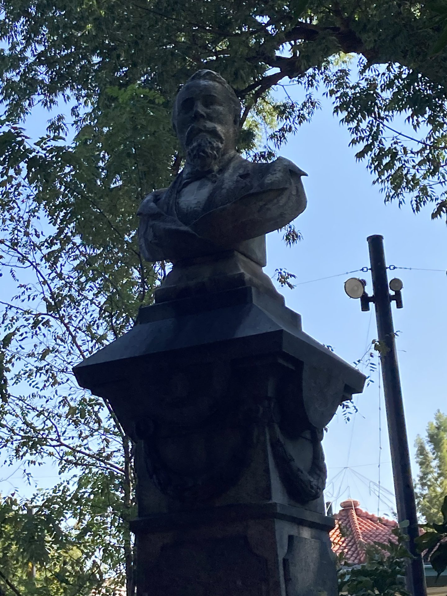 Büste eines Mannes auf einem Pfeiler, umgeben von Bäumen unter klarem Himmel.