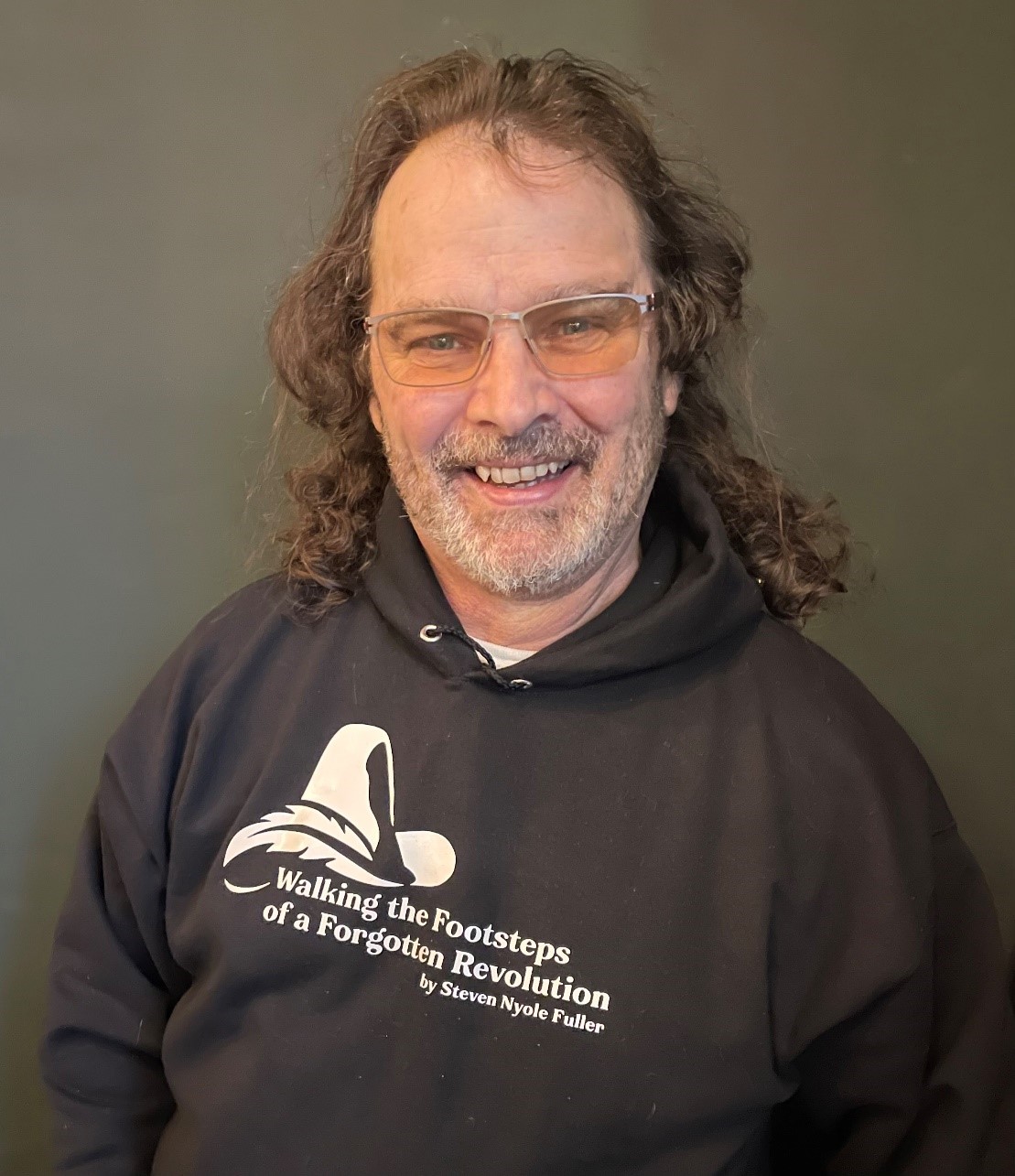 Mann mit langen Haaren und Brille lächelt im schwarzen Hoodie mit Aufdruck. Hintergrund in sanften Farben.