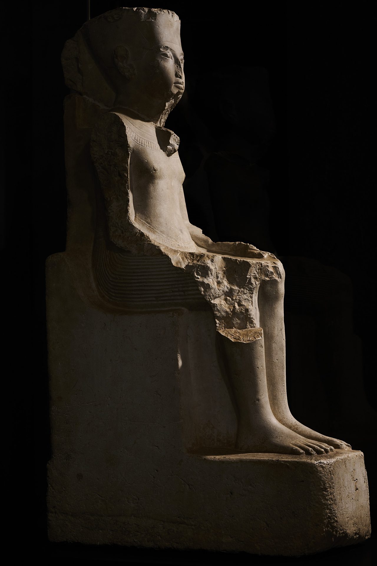 Antike Statue einer sitzenden Figur, mit erkennbaren Details und Beschädigungen. Dramatische Beleuchtung vor dunklem Hintergrund.