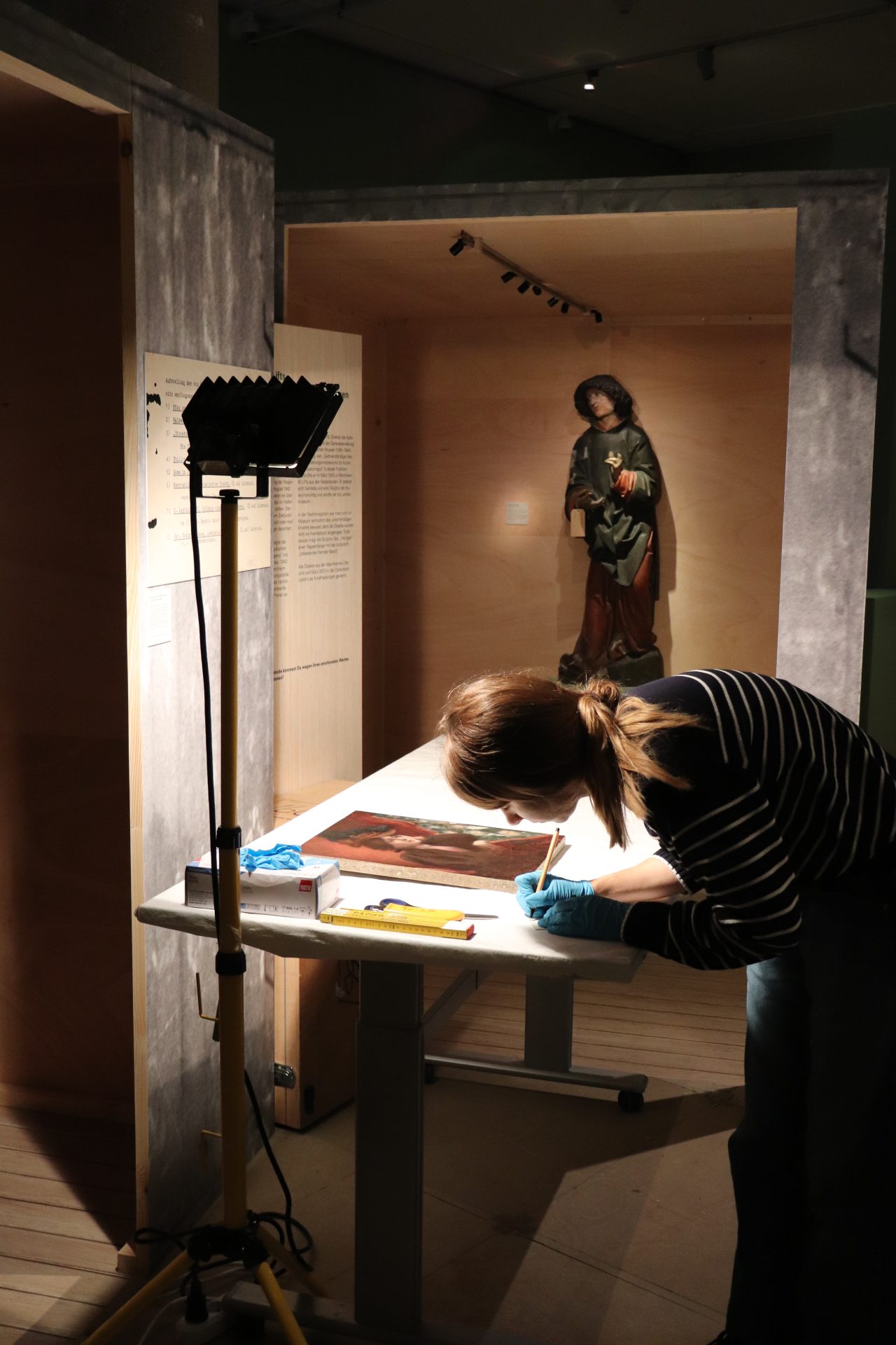 Konservatorin protokolliert ein Kunstwerk in einem beleuchteten Ausstellungsraum, im Hintergrund eine Statue.