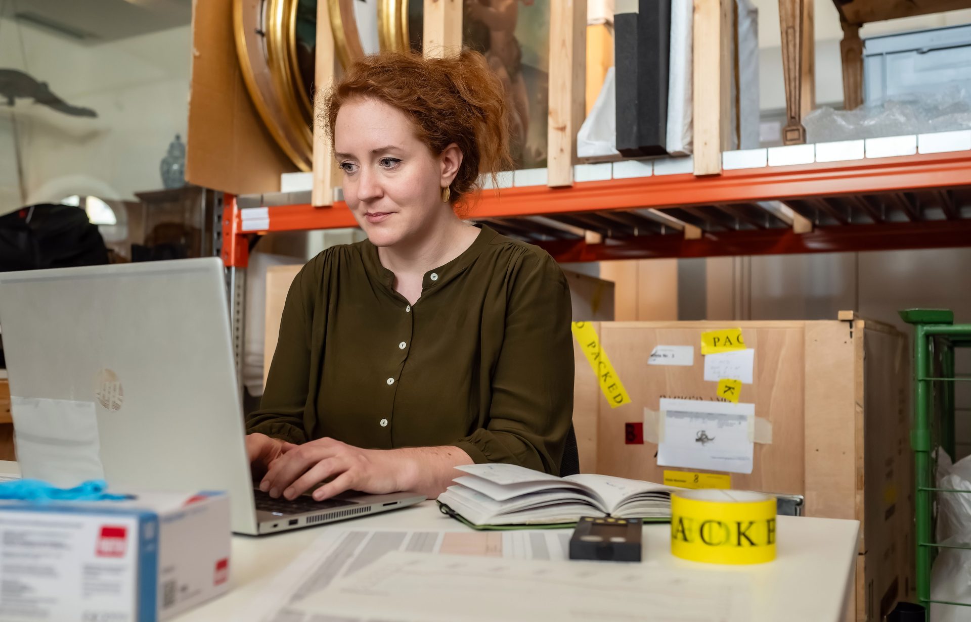 Frau mit roten Haaren sitzt an einem Laptop in einem Raum voller Kisten und Notizen.