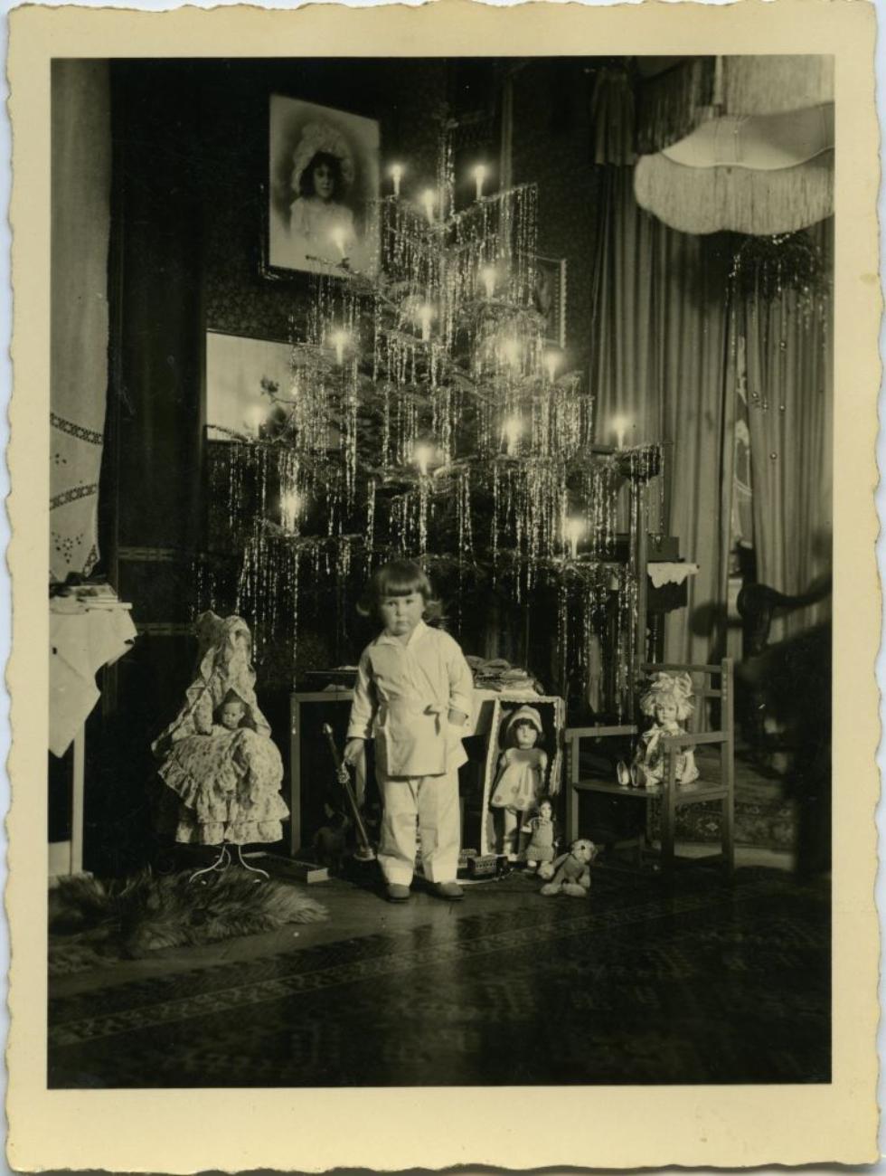 Ein Kind steht vor einem festlich geschmückten Weihnachtsbaum mit Spielzeug und Puppen in nostalgischer Stimmung.