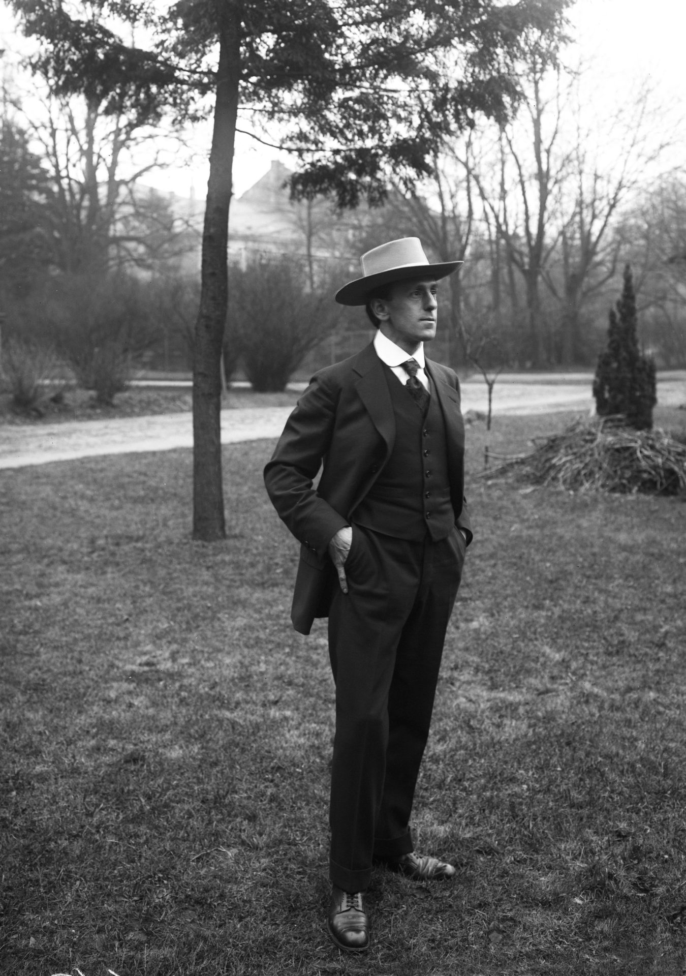 Ein Mann in Anzug und Hut steht nachdenklich im Garten, umgeben von Bäumen und Sträuchern. Schwarzweißaufnahme.