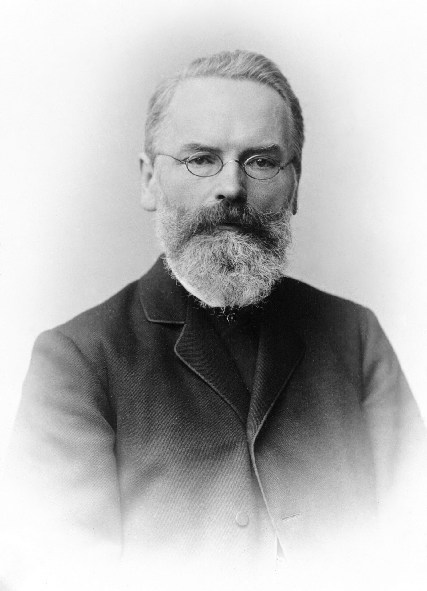 Porträt eines Mannes mit Brille, Bart und Anzug. Sein ernsthafter Ausdruck verleiht ihm eine historische Anmutung.