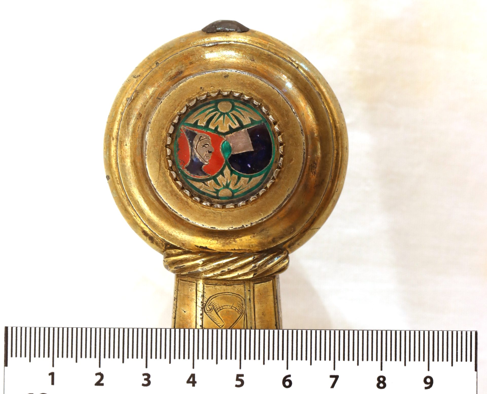 Zierlicher goldener Gegenstand mit buntem Emblem einer stilisierten Figur und ornamentalen Details.