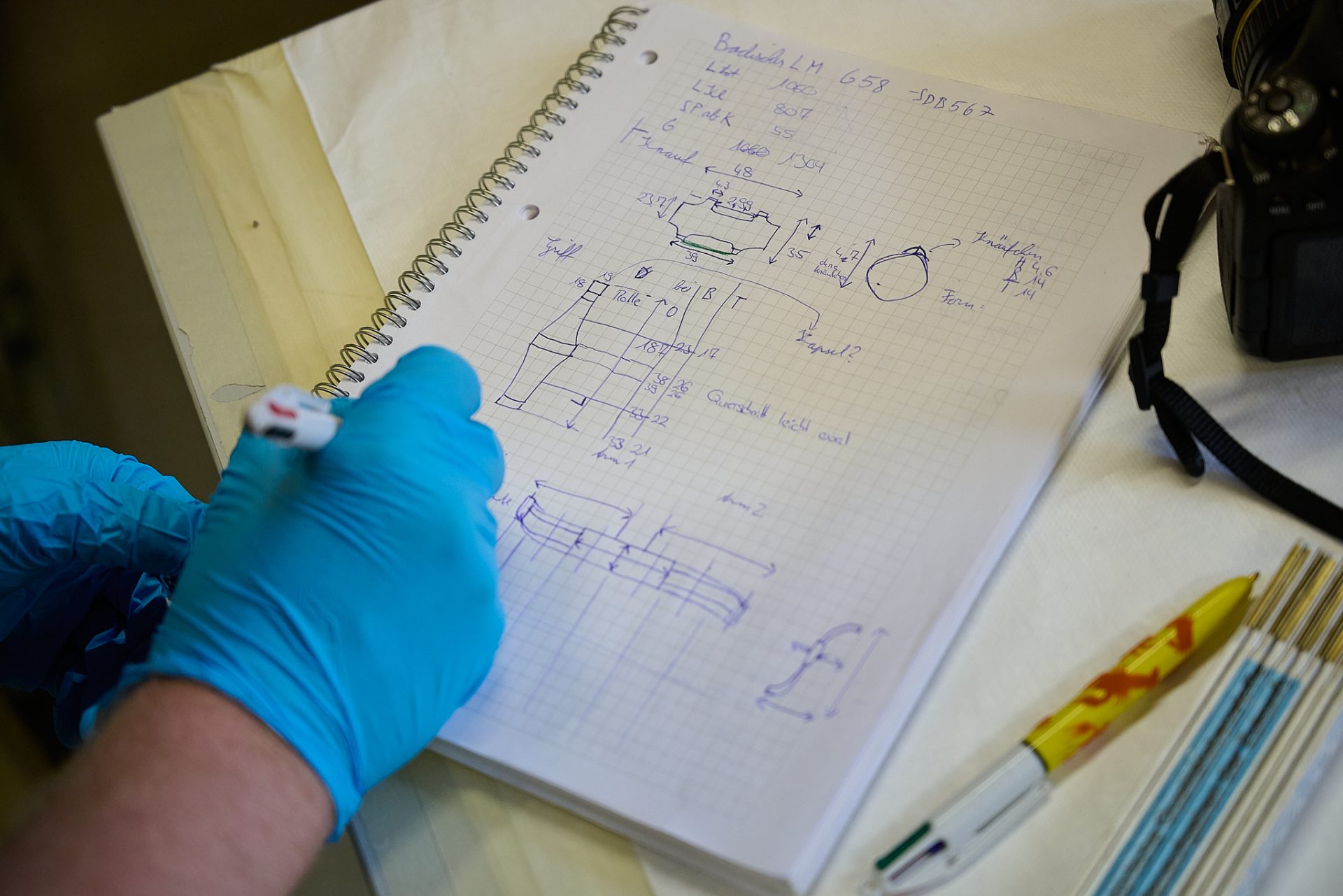 Notizbuch mit technischen Zeichnungen, jemand schreibt mit einem Marker, umgeben von Stiften und einer Kamera.