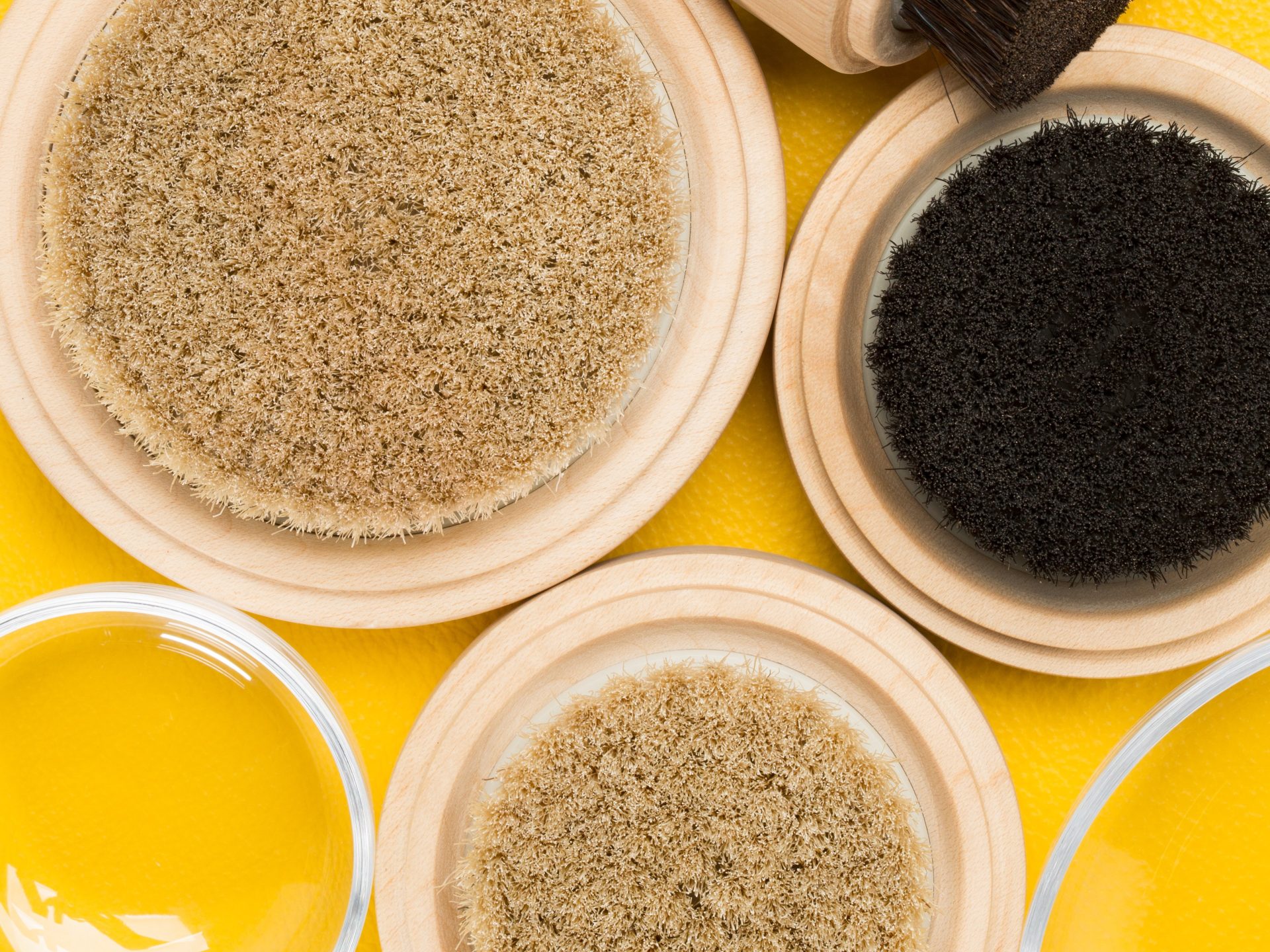 Drei runde Schalen mit verschiedenen Materialien in neutralen Farben auf gelbem Hintergrund, mit Pinsel und Glaskappe.