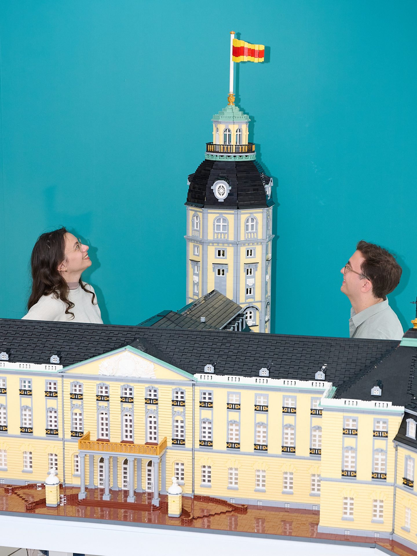 Zwei Personen betrachten ein detailliertes Lego-Modell eines Gebäudes mit einer Fahne vor einer blauen Wand.