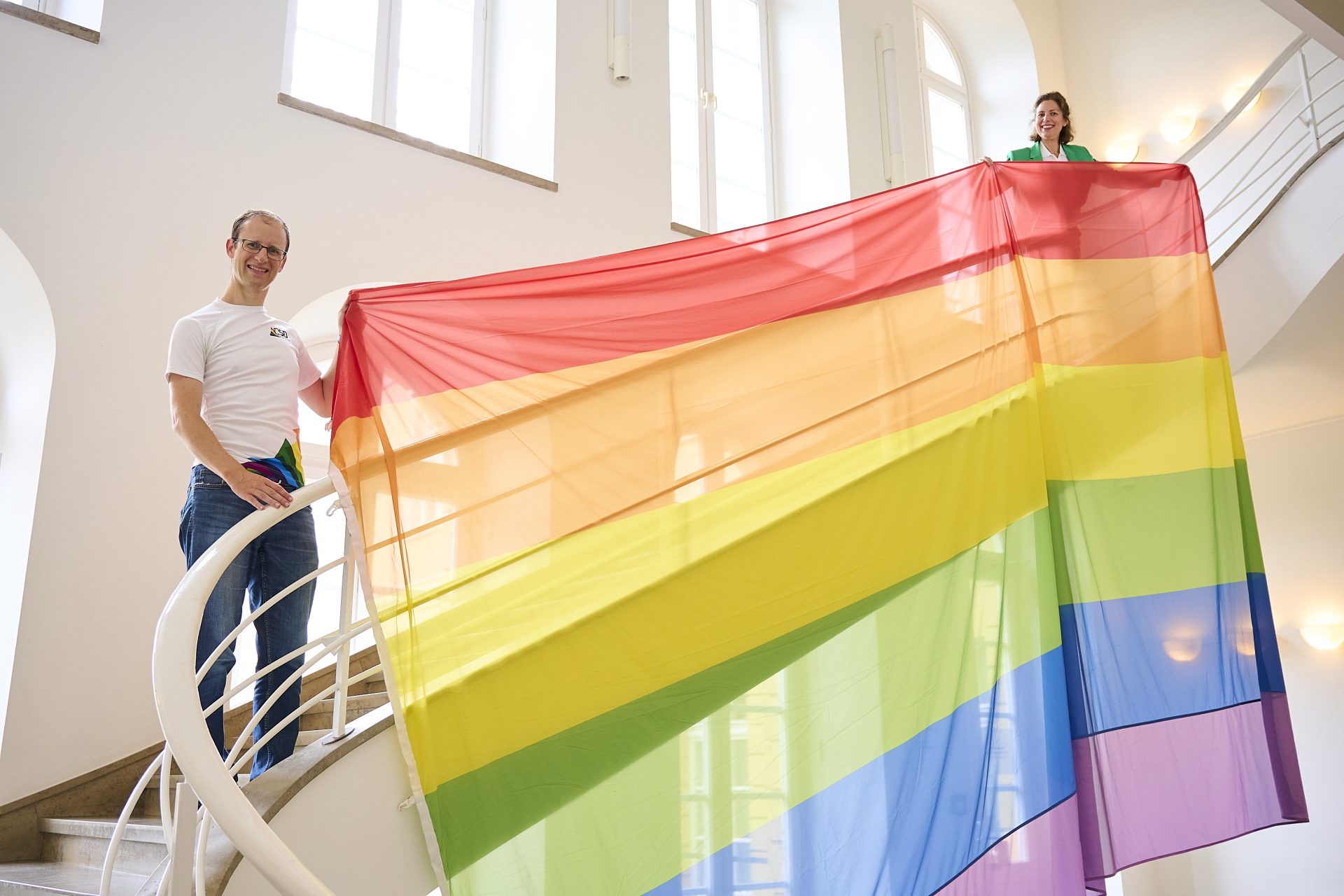 Zwei Personen präsentieren stolz eine große Regenbogenflagge in einem hellen Treppenhaus.
