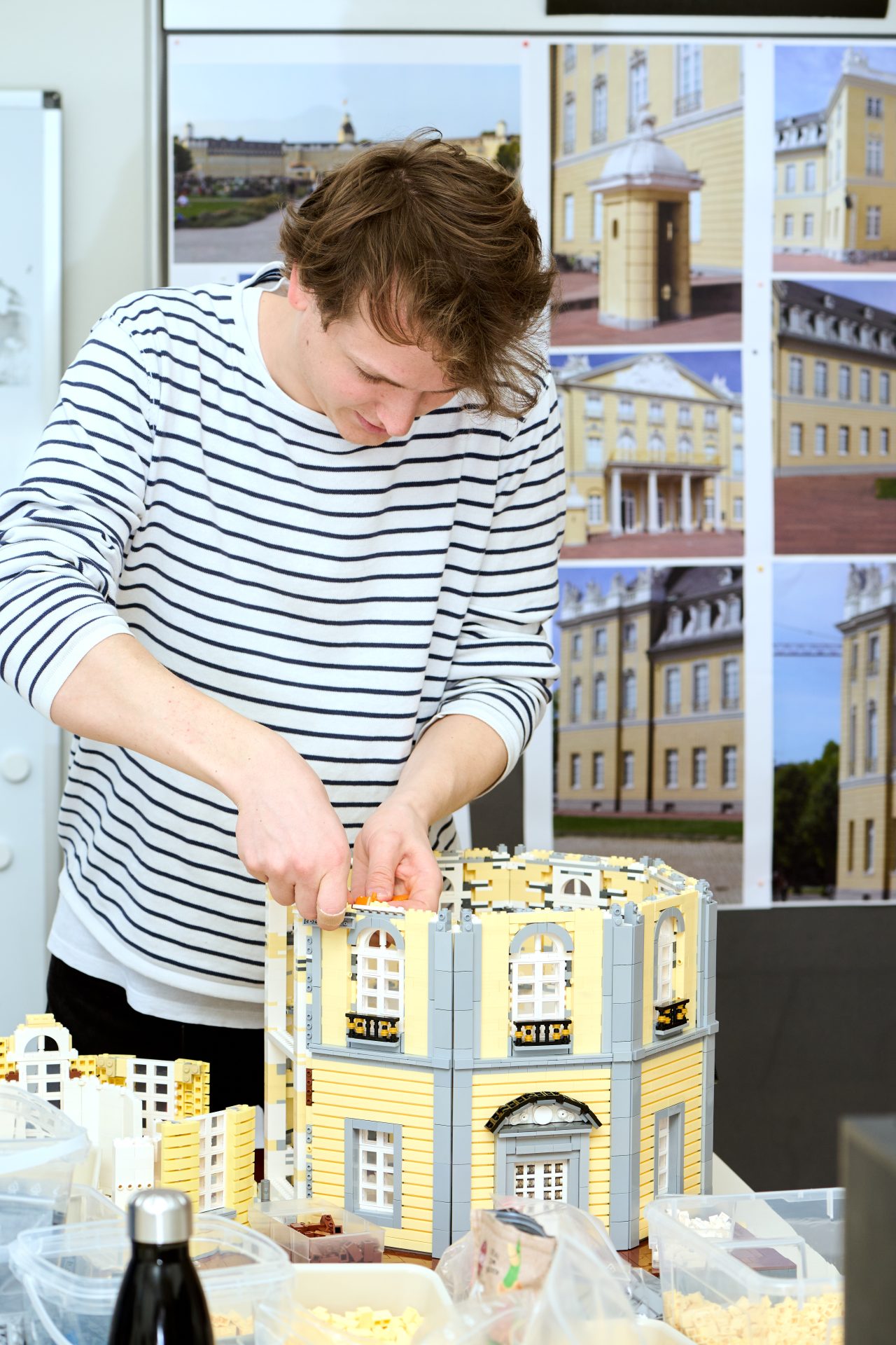 Junger Mann baut ein LEGO-Modellhaus auf einem Tisch mit Bauteilen, umgeben von Bildern des Originals.