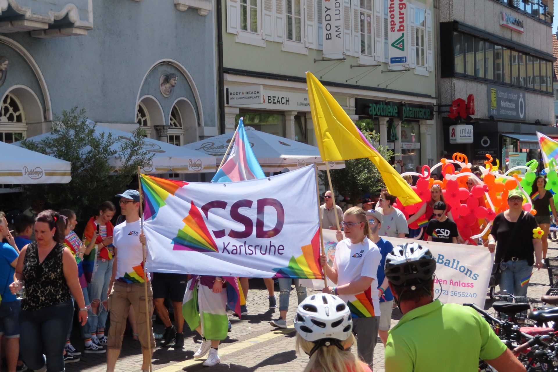 CSD Karlsruhe Parade mit bunten Fahnen und Luftballons in einer sonnigen Stadtstraße.